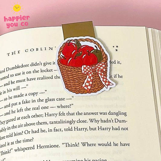 A magnetic bookmark of a red apple basket with a red-and-white checkered bow, placed on an open book page. The sticker has a brown kraft-look top backing and a white border.