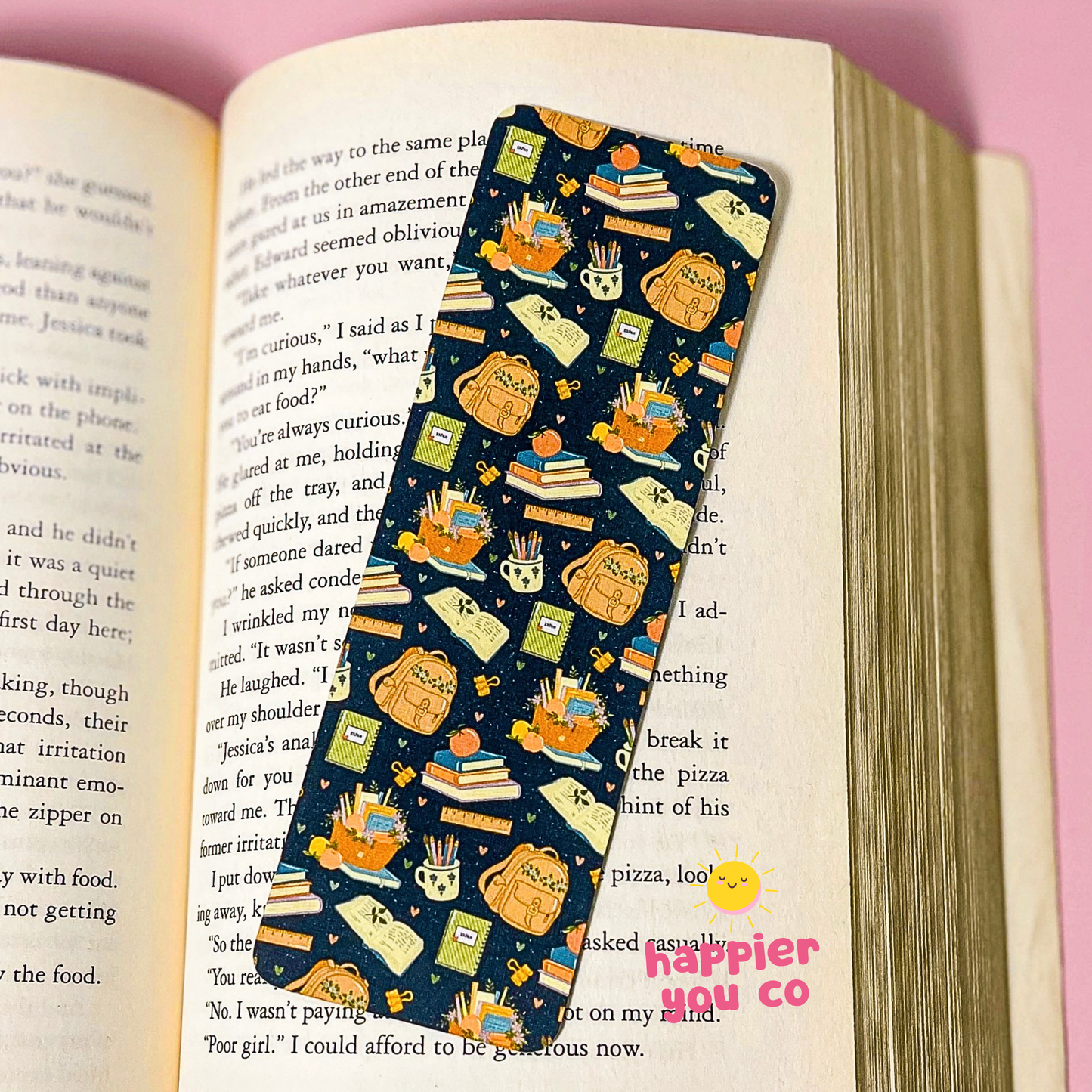A long, narrow bookmark with a navy blue background and a playful pattern of cartoon cats, books, and reading accessories, resting on an open book with a pink background.