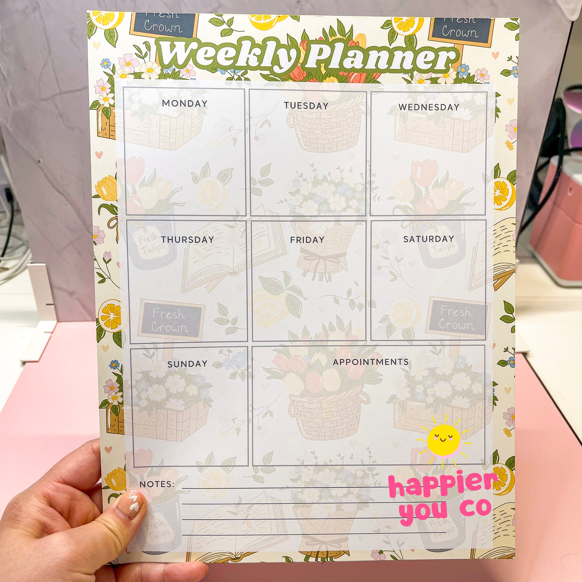 Weekly planner with floral design held by a hand, featuring the brand 'Happier You Co'.