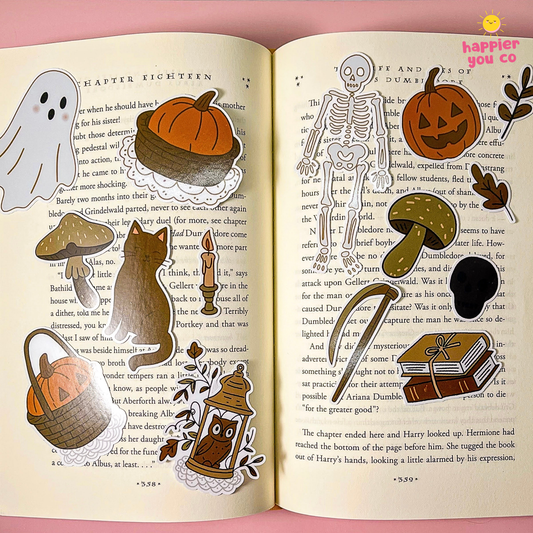 A set of Halloween-themed sticker cutouts arranged on an open book. The stickers include a ghost, pumpkin, mushrooms, a cat, a witch, a skeleton, a basket, a book, and leaves, all in a coordinated autumn color palette.