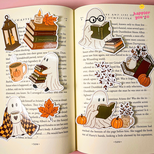 An open book spread styled as a Halloween-themed ghost and bookish collage, featuring stickers of white ghosts, pumpkins, autumn leaves, old books, and a lantern over printed text pages. The pink background around the open book has a small 'Happy Ghosts' logo in the top right.