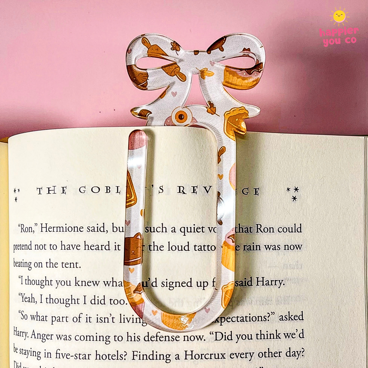A decorative paper bookmark shaped like a bow and arrow is clipped onto an open book. The bookmark has a white base with orange, yellow, and brown patterns, set against a pink background.