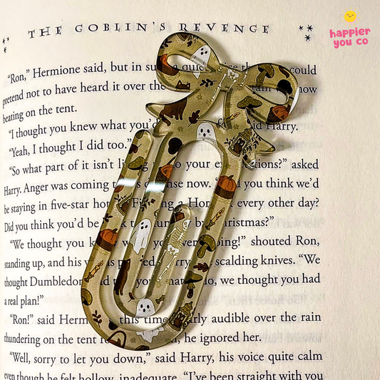 A decorative paperclip bookmark featuring a Halloween theme with ghosts, pumpkins, and spider motifs, resting on an open book page.