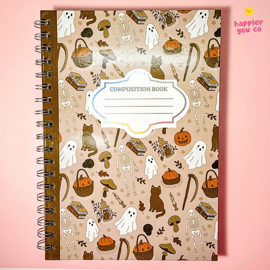 A spiral-bound composition notebook with a beige cover decorated with a Halloween pattern of ghosts, skeletons, pumpkins, mushrooms, and cats. The notebook is photographed flat against a solid pink background, with a small 'happier you co' logo in the top right corner.