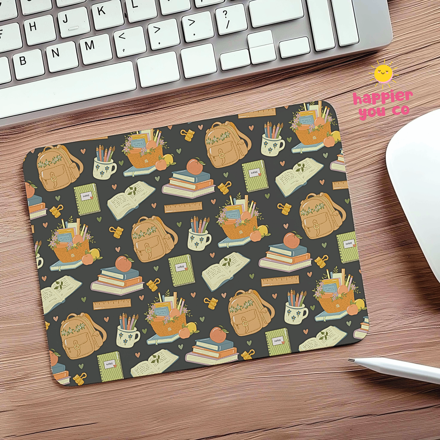 A rectangular desk mat with a repeating autumn-themed school supplies pattern, placed on a wooden desk next to a keyboard, mouse, and stylus. The pattern features tan backpacks, books, pencils, and cups on a dark gray background.