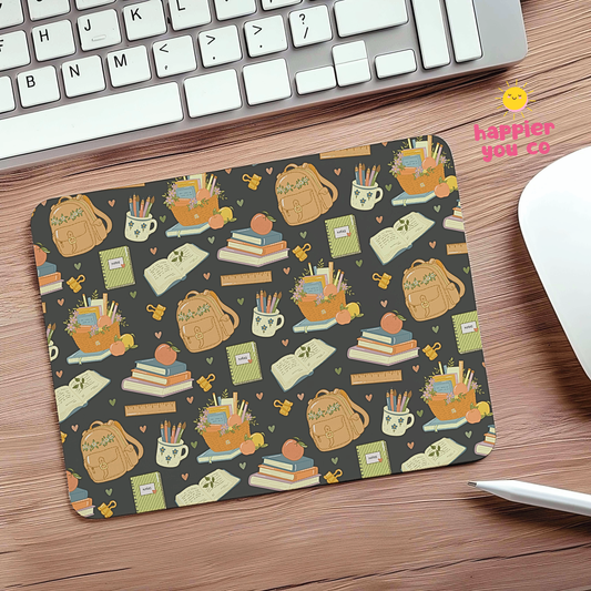 A rectangular desk mat with a repeating autumn-themed school supplies pattern, placed on a wooden desk next to a keyboard, mouse, and stylus. The pattern features tan backpacks, books, pencils, and cups on a dark gray background.