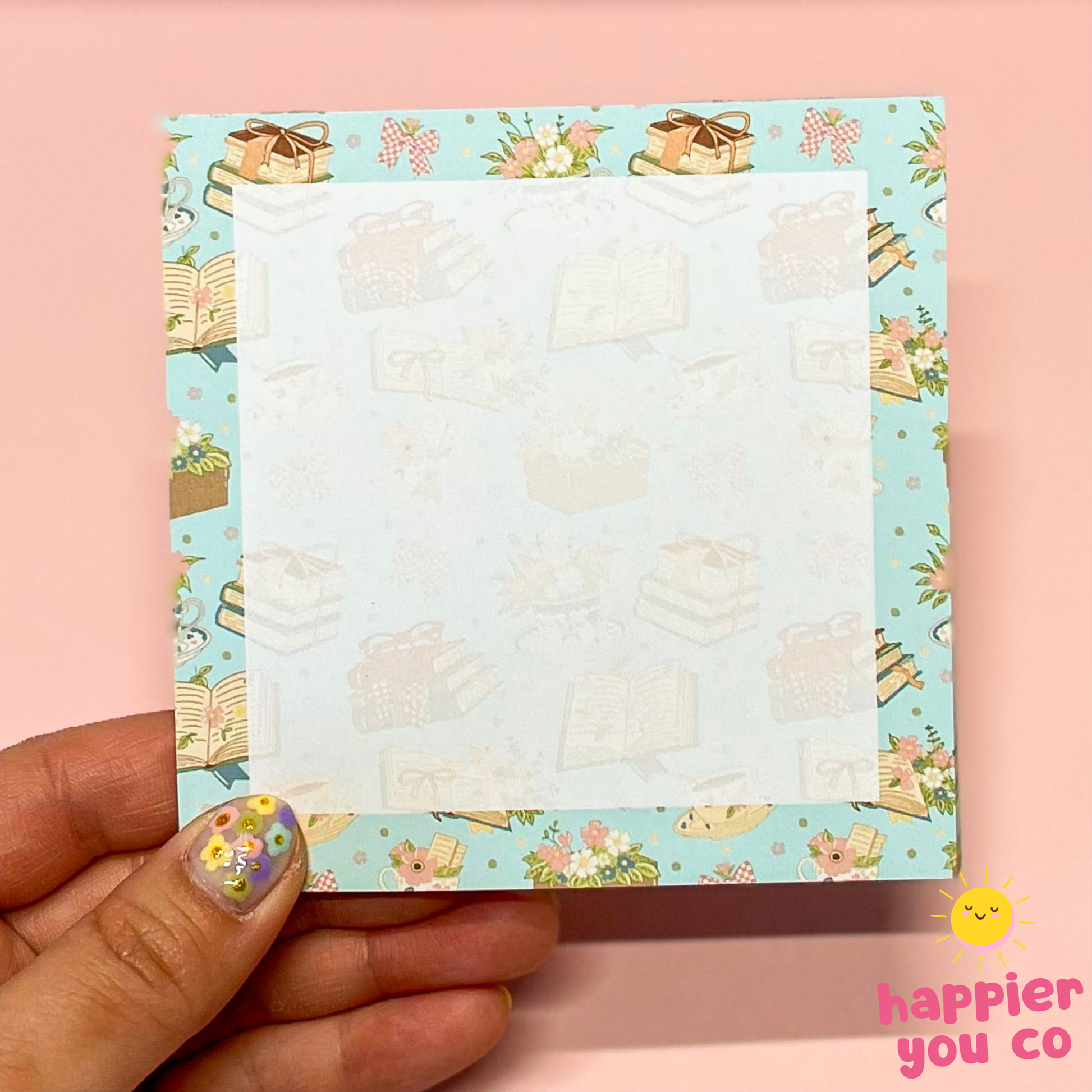 Hand holding a small notepad with a floral and book design on a pink background