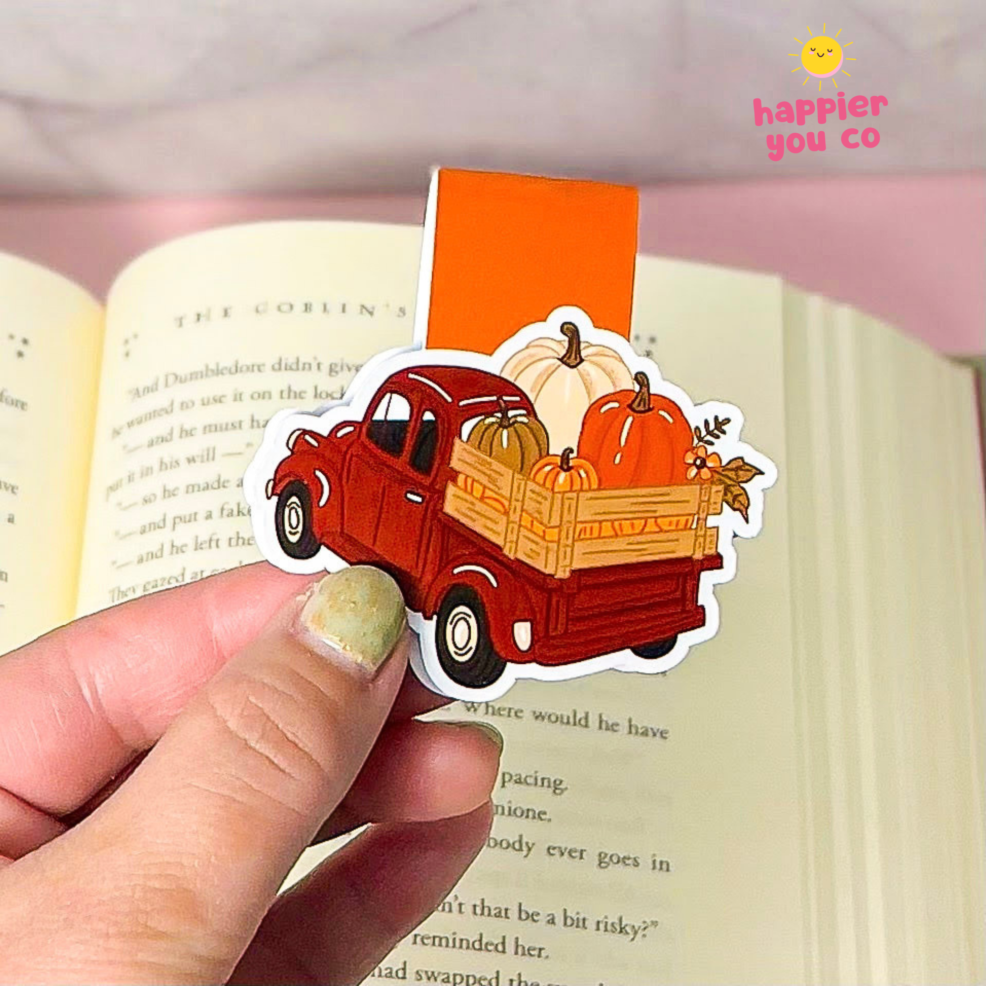 A hand holds a die-cut sticker shaped like a red vintage pickup truck carrying pumpkins in a wooden bed, positioned over an open book. The sticker has a white border and a small orange brand mark reading 'happier you co' in the top right corner.