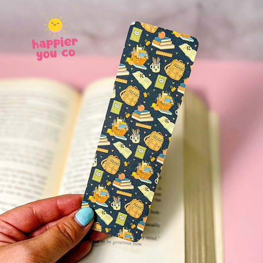 A hand with light blue nail polish holds a long rectangular bookmark or card featuring a navy blue background with a repeating pattern of cute illustrated books, books on shelves, and small objects in yellow, cream, and green. An open book and a pink surface are blurred in the background, and the text 'happier you co' with a yellow smiley appears in the upper left.