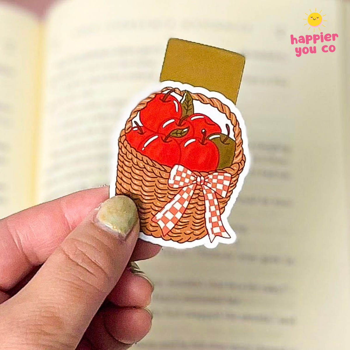 A hand holds a magnetic bookmark shaped like a woven basket filled with red apples and tied with a red-and-white checkered bow. The sticker is photographed over an open book, with a small 'happier you co' logo in the top right corner.