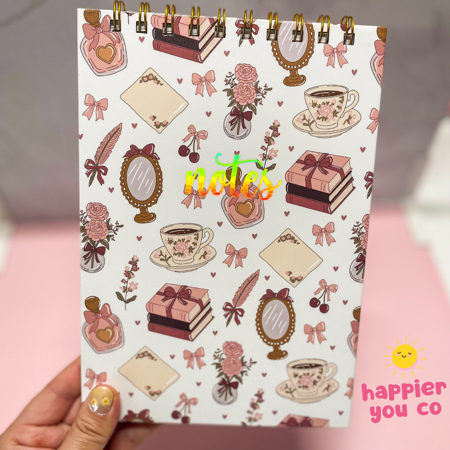 Notebook with decorative cover featuring illustrations of tea cups, flowers, and bows held by a hand.