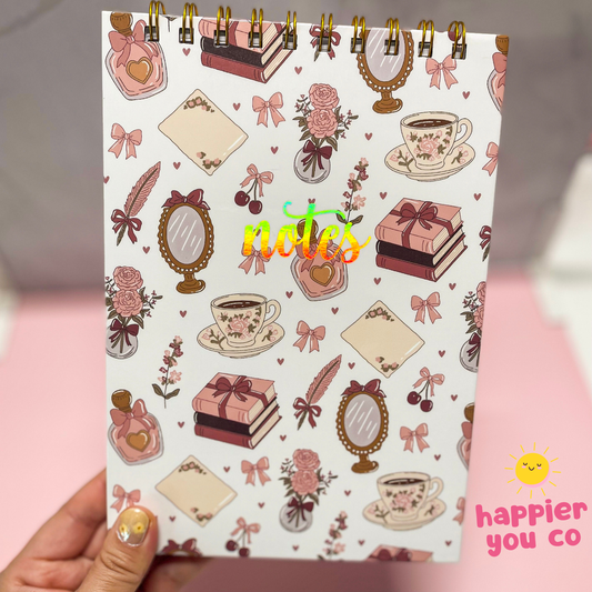 Notebook with decorative cover featuring illustrations of tea cups, flowers, and bows held by a hand.