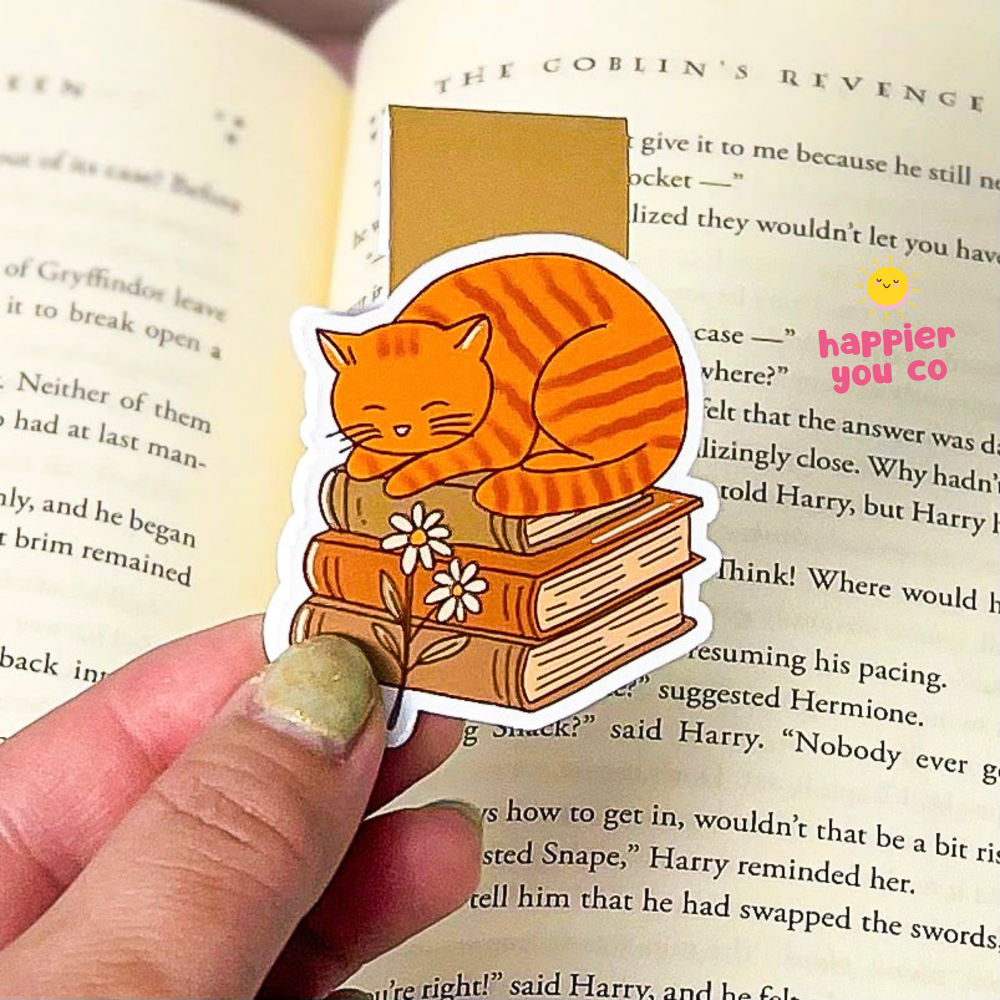 A magnetic bookmark of an orange tabby cat sleeping on stacked books with small white flowers, held by a hand in front of an open book.