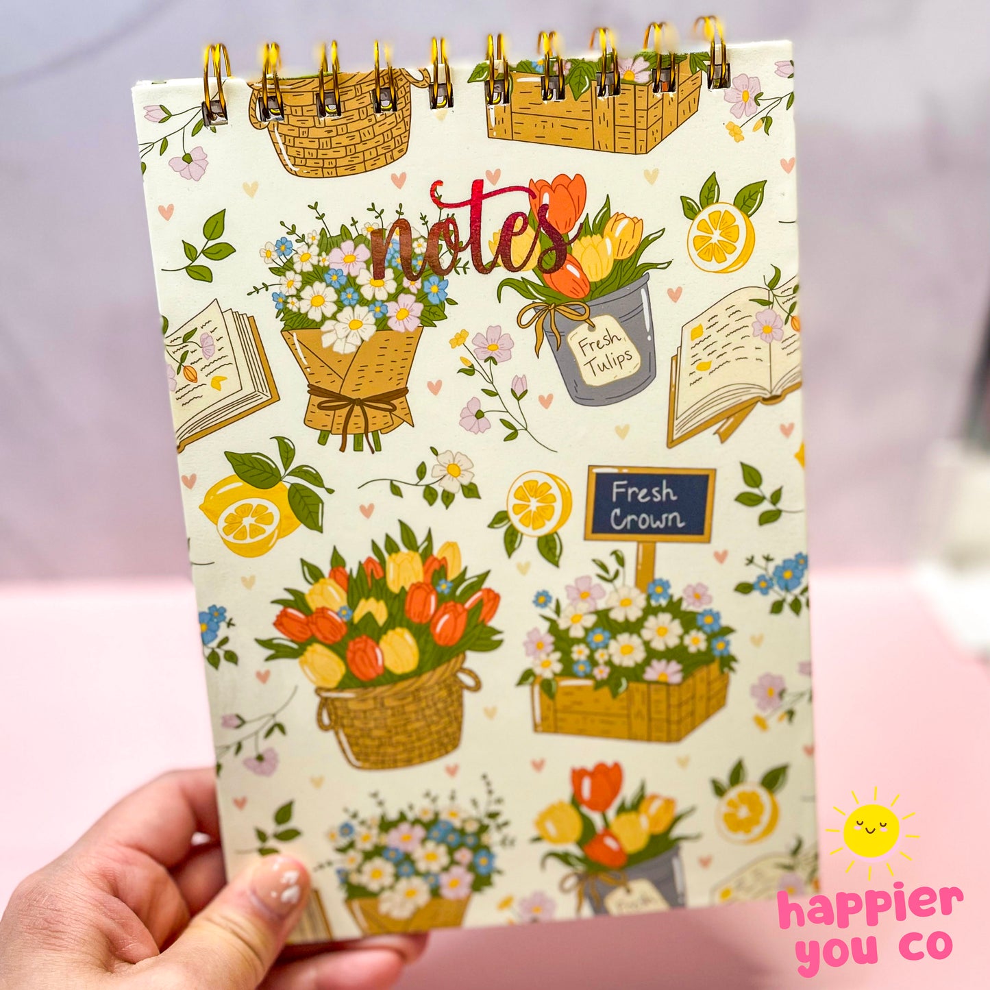 Notebook with floral design and 'notes' text held by a hand on a light pink background.