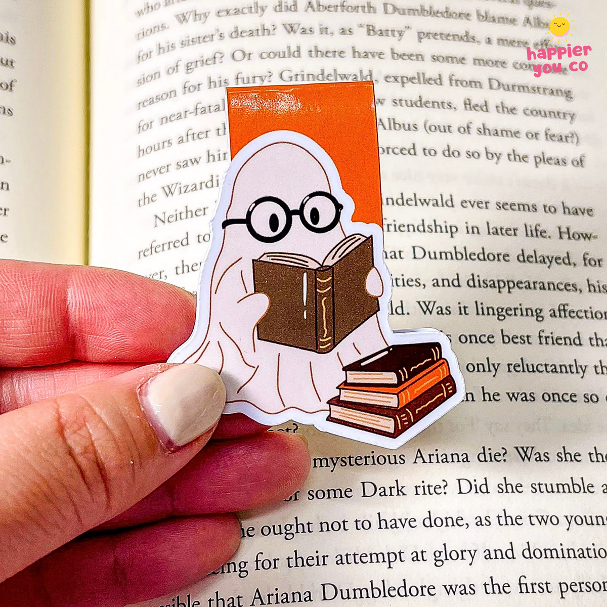 A hand holds a die-cut bookmark shaped like a ghost reading books, placed over an open book page. The sticker has a white ghost body, round black glasses, and brown books, with a bright orange rectangular accent behind the ghost.