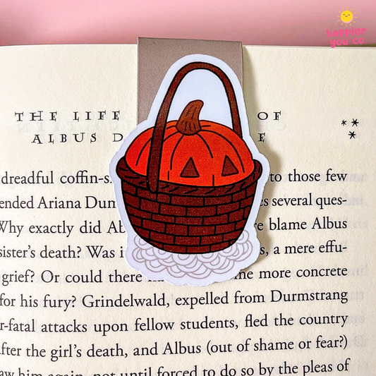 A die-cut sticker of a pumpkin-shaped basket bookmark resting on an open book page. The sticker features a red-orange pumpkin lid with triangular window details, a brown woven basket, and a white cloud-like base.