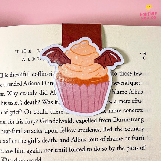 A die-cut bookmark shaped like a cupcake with bat wings, placed on an open book page. The sticker has a pink wrapper, light orange frosting, and dark red wings.