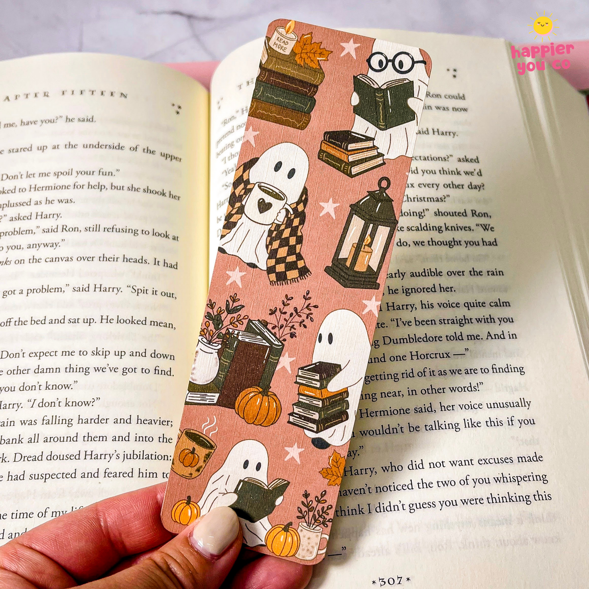 A pink autumn-themed bookmark with illustrations of ghosts, books, pumpkins, and stars is held in a hand and placed across open book pages.