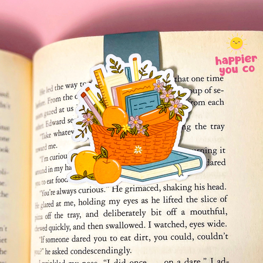 A magnetic bookmark featuring an orange woven basket filled with books, pencils, flowers, and oranges, placed on an open book page. The sticker has a white border and is shown against a soft pink background.