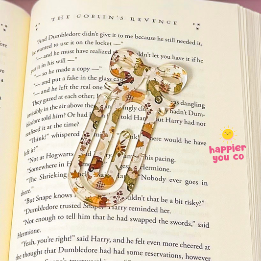 A decorative paperclip bookmark styled like a book page with Harry Potter references, featuring a cute cartoon character and autumn leaves, resting on an open book.