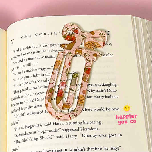 A decorative paperclip bookmark with a pink and orange floral pattern and small green leaves, resting on open book pages. The bookmark features a large bow on top with the words 'happier you co' printed in pink next to a small yellow sun graphic.
