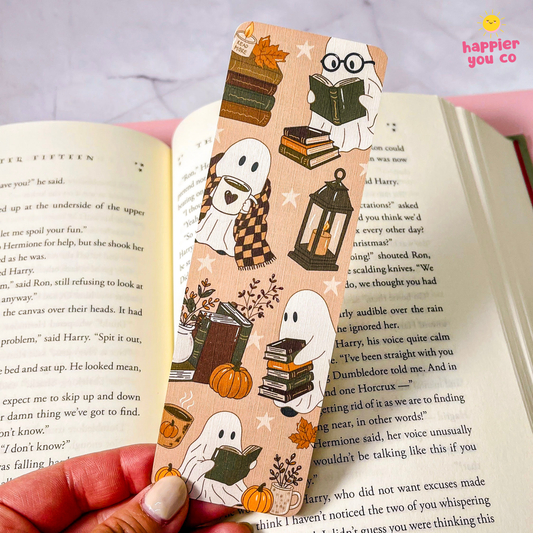 A hand holds a decorative bookmark with a fall ghost-themed illustration over an open book. The bookmark features cartoon ghosts, stacked books, a lantern, pumpkins, stars, and autumn leaves on a peach background.