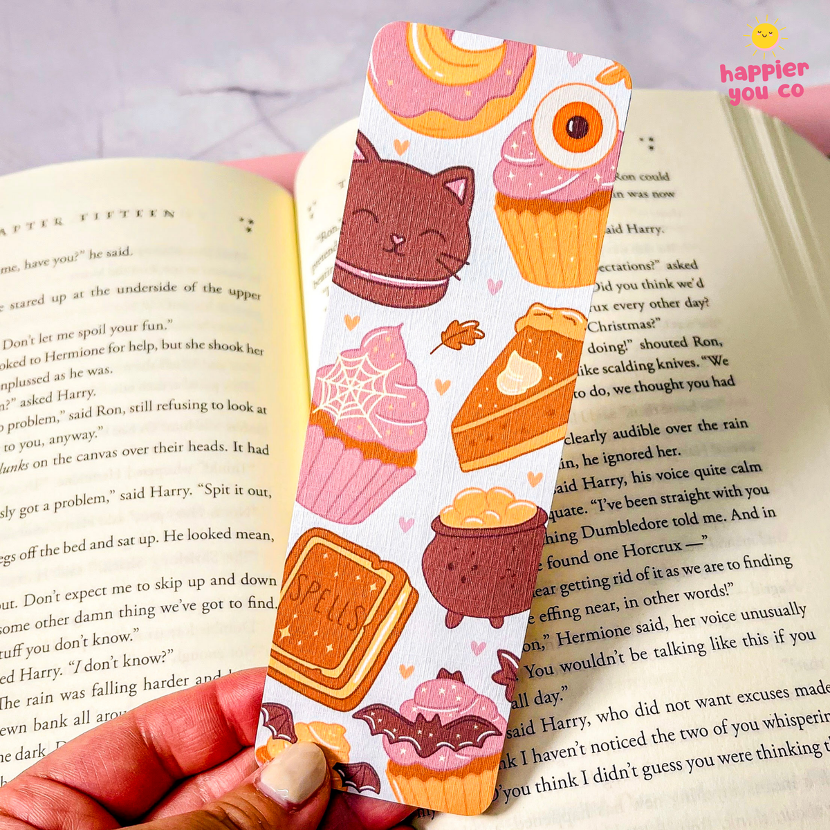A hand holds a pastel-colored bookmark shaped like a book page, decorated with cute illustrations of cupcakes, a cat, a spiderweb, and a cookie. The bookmark is shown partially inserted into an open book.