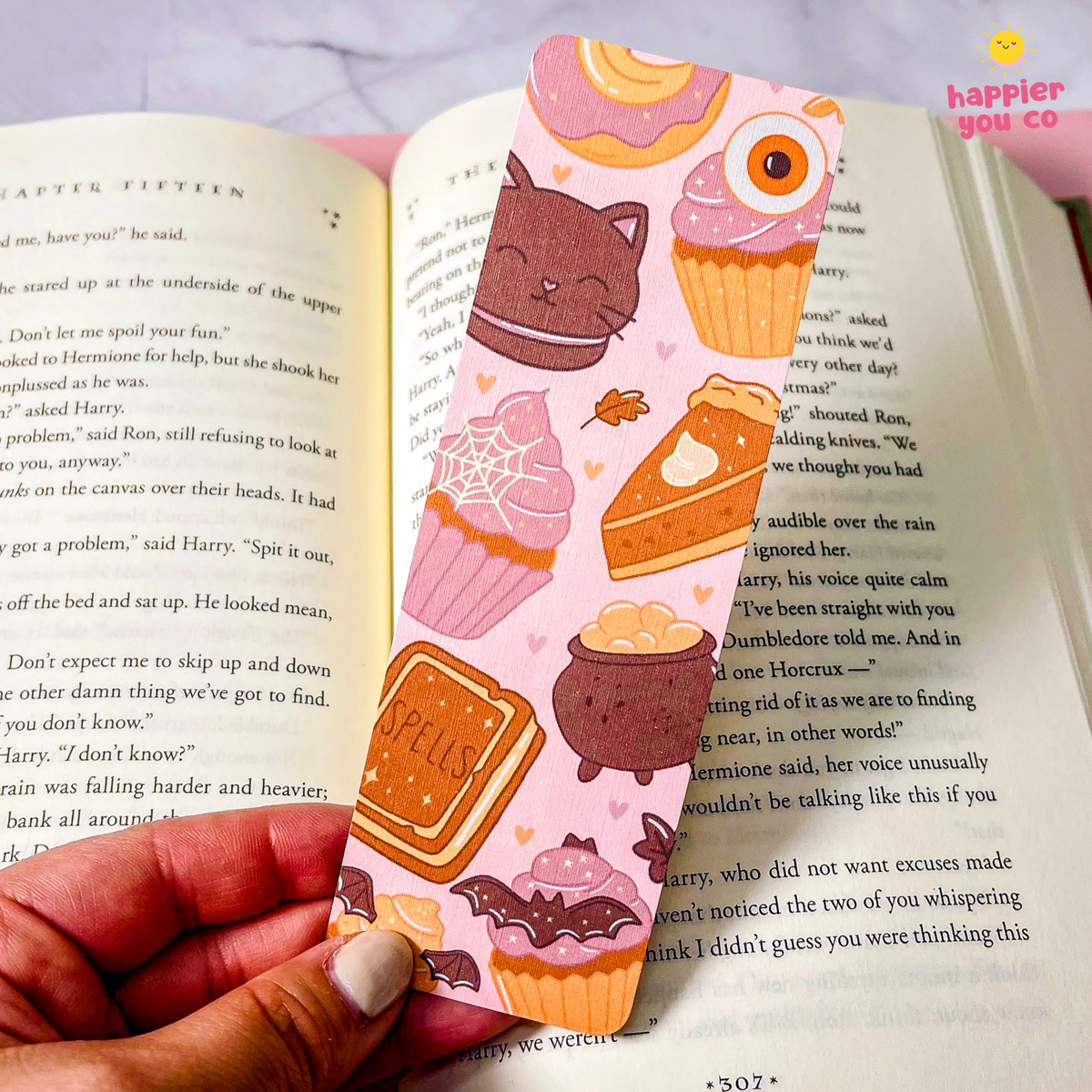 A close-up of a hand holding a pink and orange illustrated bookmark or sticker sheet over an open book. The sheet features cartoon cupcakes, a cat, a slice of cake, and a chocolate bar with the word 'SPELLS'.