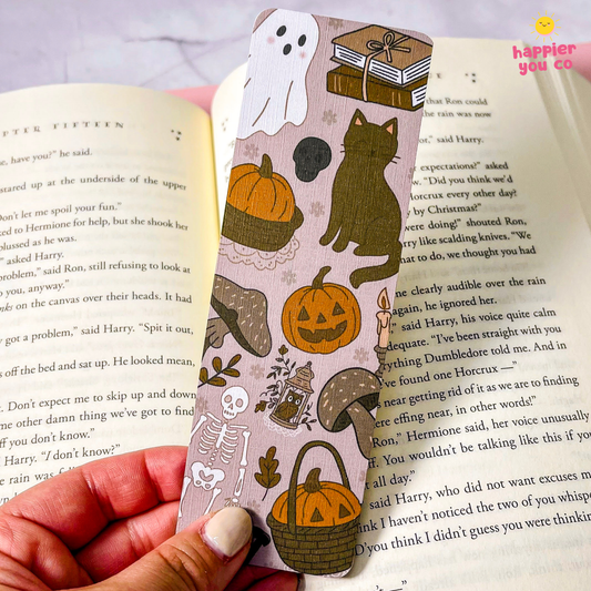 A hand holds a pink bookmark decorated with Halloween illustrations, including pumpkins, a cat, a ghost, a skeleton, and a fox, positioned over an open book.