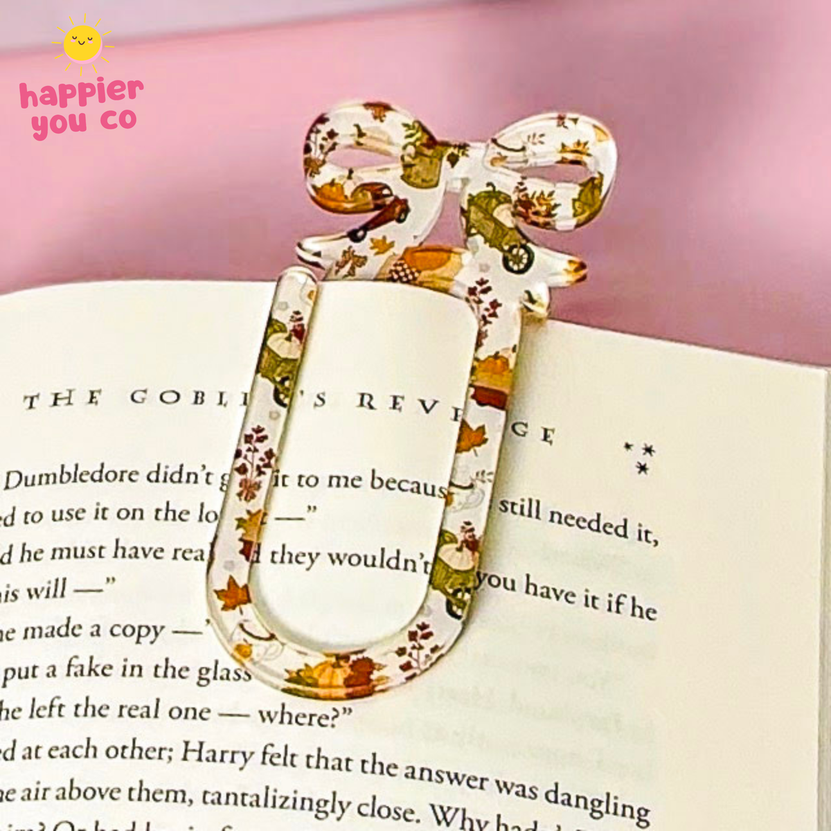 A decorative paperclip-shaped bookmark with a bow at the top, printed with a fall-themed pattern of orange leaves, pumpkins, and autumn foliage on a cream background. It is shown resting on an open book with a pink background.