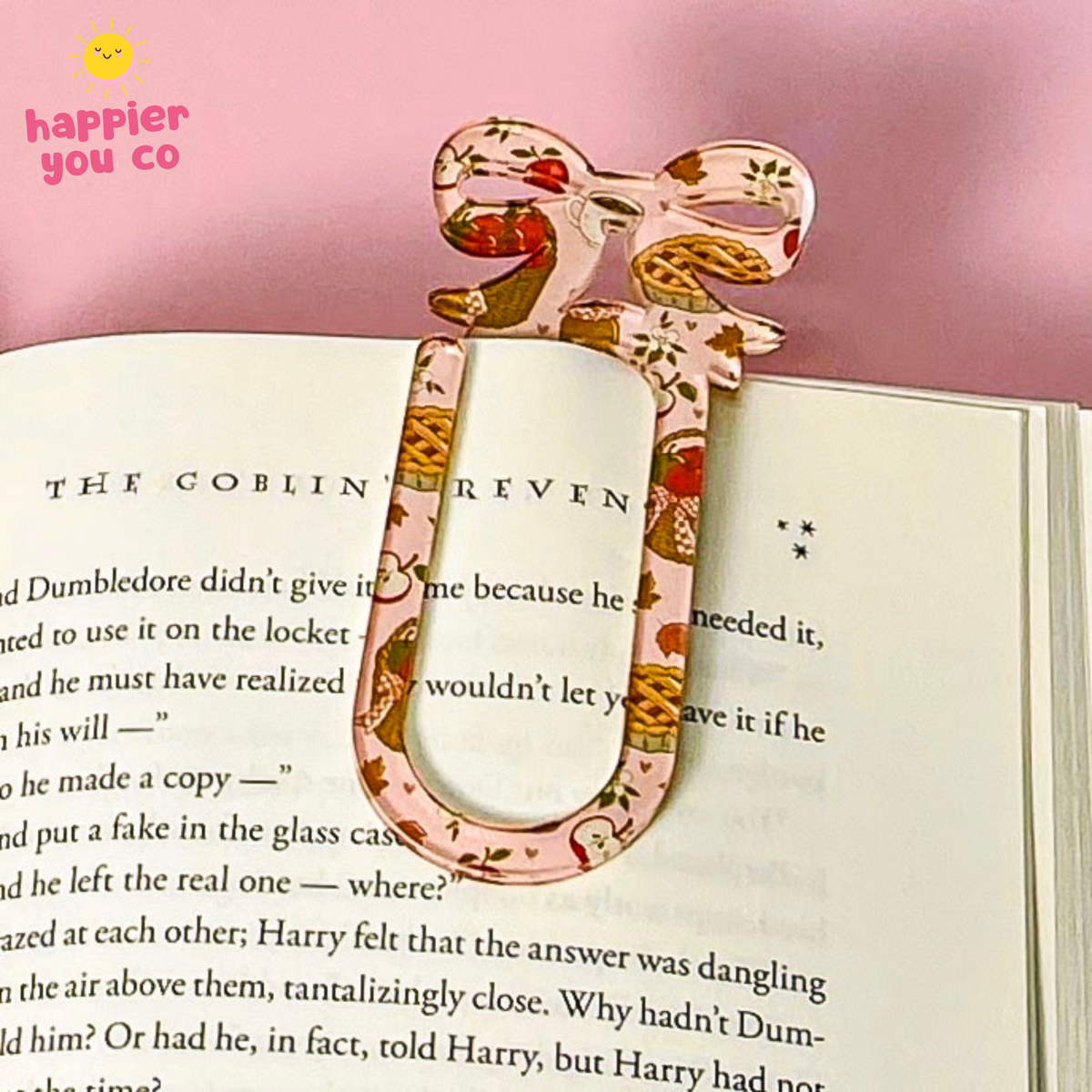 A decorative U-shaped book bookmark shaped like a bow, featuring a pink and white floral pattern with small red and gold accents, resting on an open book against a pink background. A 'happier you co' logo is visible in the top left corner.