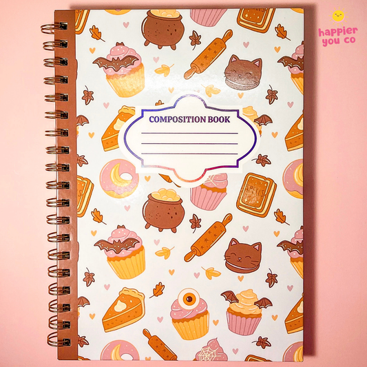 A spiral-bound composition book with a white cover decorated with cute autumn-themed illustrations including cupcakes, donuts, pies, cauldrons, rolling pins, bats, and leaves. The center features a purple-bordered label that reads 'COMPOSITION BOOK'.