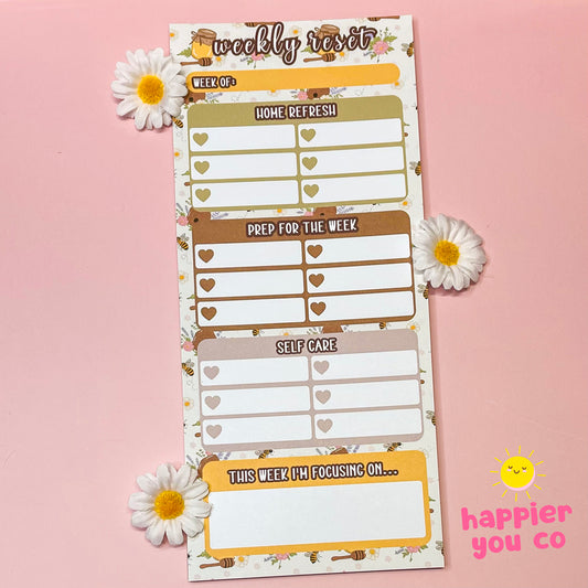 Weekly reset planner pad with floral bee design on a pink background, featuring the brand 'happier you co'.