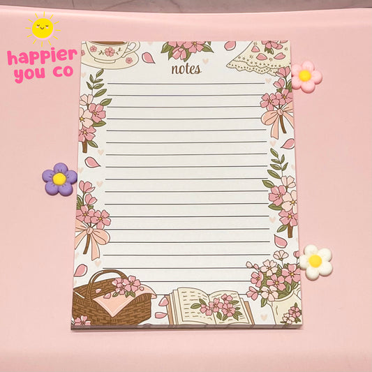 Notepad with decorative floral border on a pink background, featuring the brand 'happier you co'.