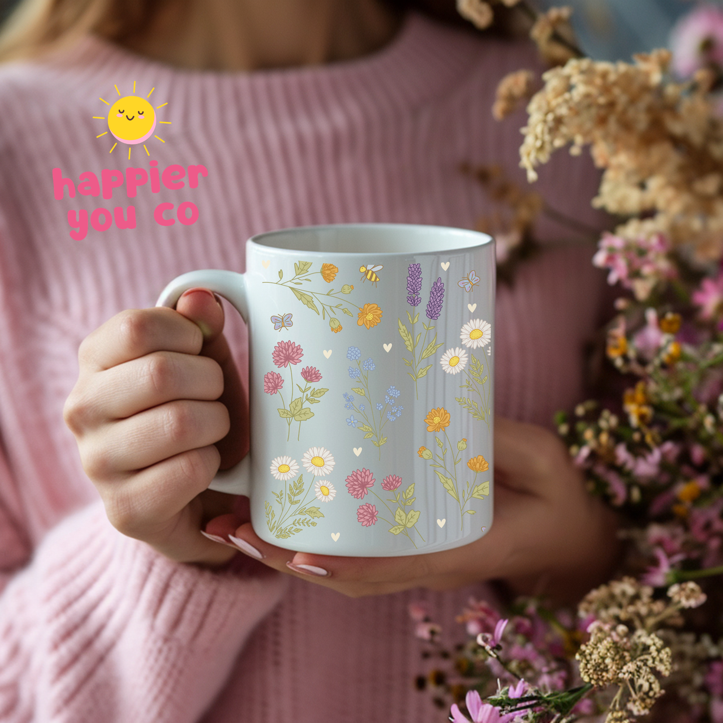 Person holding a mug with floral design, surrounded by flowers, with 'happier you co' logo.