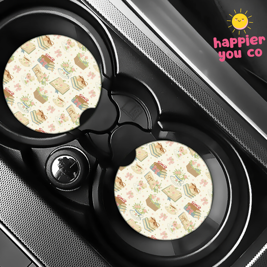 Two car coasters with a pattern of books and flowers on a cup holder tray, branded 'happier you co'.
