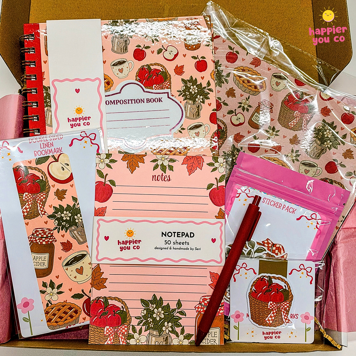 A stationery gift set from happier you co. featuring a pink apple-themed composition book, a lined notepad, a sticker pack, and other matching paper goods arranged in a box with a red pen.