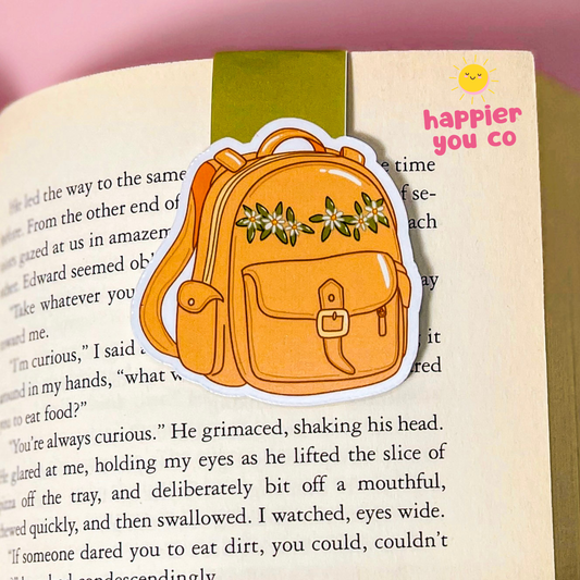 A magnetic bookmark of an orange backpack with green floral details, shown resting on an open book page next to a green bookmark. The sticker has a white border and is photographed against a soft pink background.