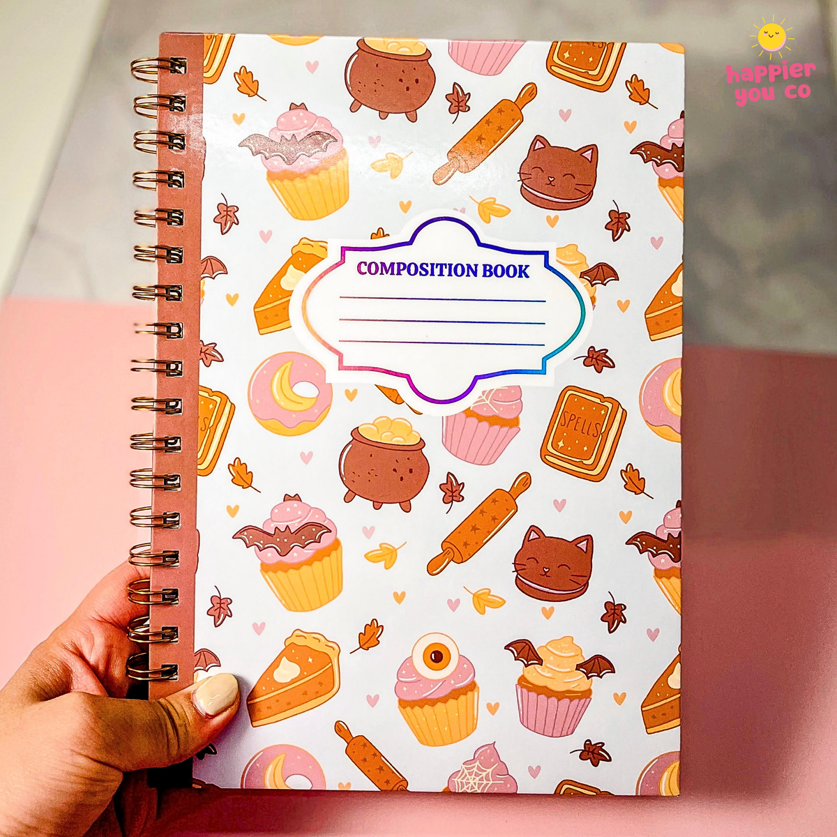 A hand holds a spiral-bound composition notebook with a white cover decorated with cute illustrations of cupcakes, cauldrons, rolling pins, cookies, and cat faces. The notebook features a blue and purple outlined label that reads 'COMPOSITION BOOK'.