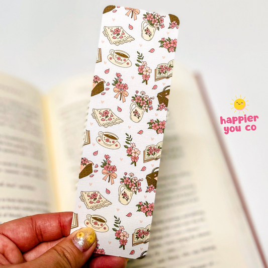 A hand holds a long, narrow bookmark or card featuring a repeating pattern of pastries, flowers, and books on a white background. An open book is blurred in the background, and a pink 'happier you co' logo watermark appears on the right.
