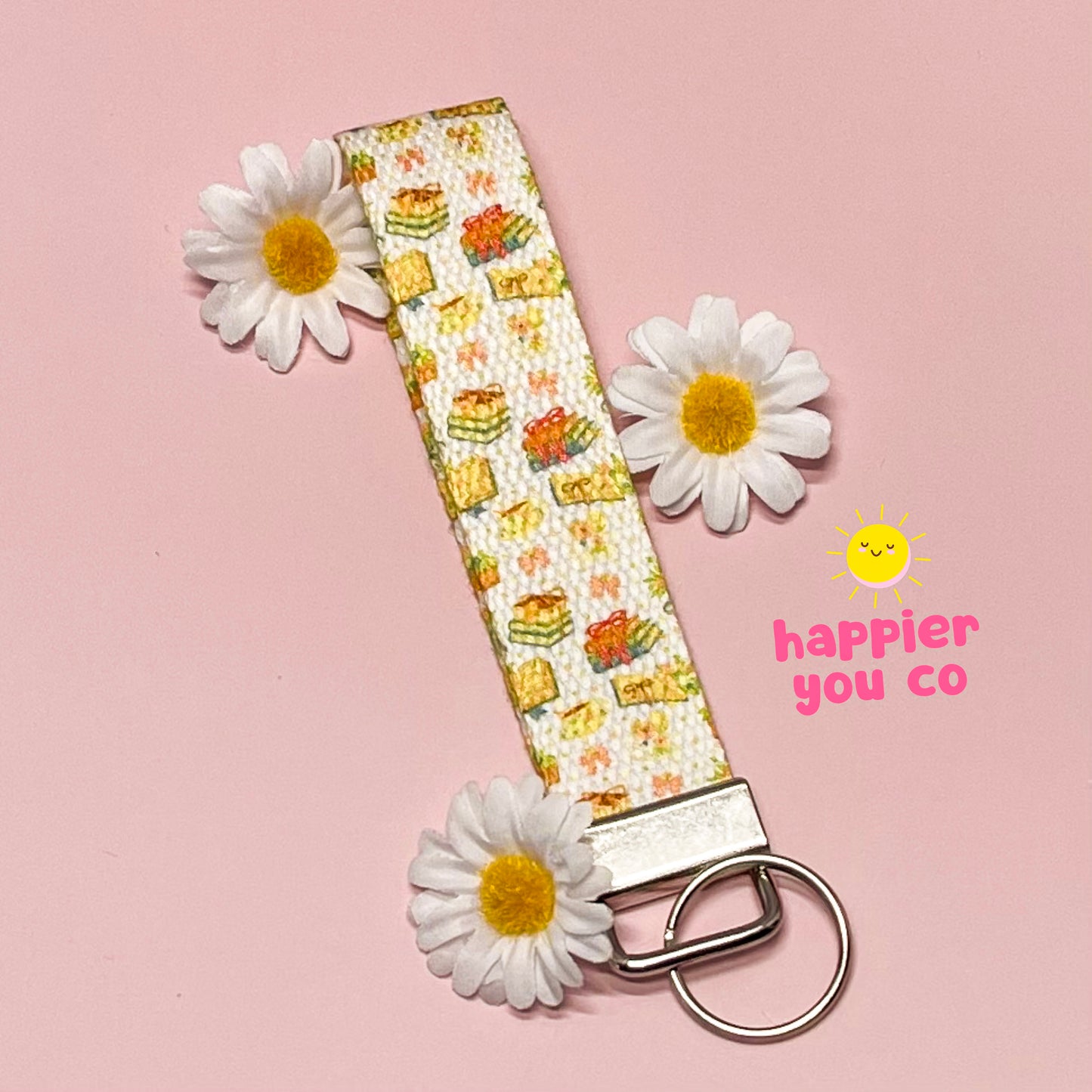 Keychain with floral pattern and daisies on a pink background, featuring the brand 'happier you co'.