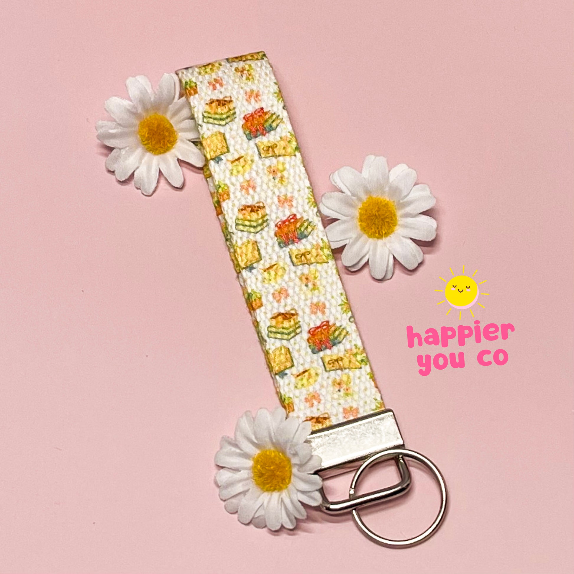 Keychain with floral pattern and daisies on a pink background, featuring the brand 'happier you co'.