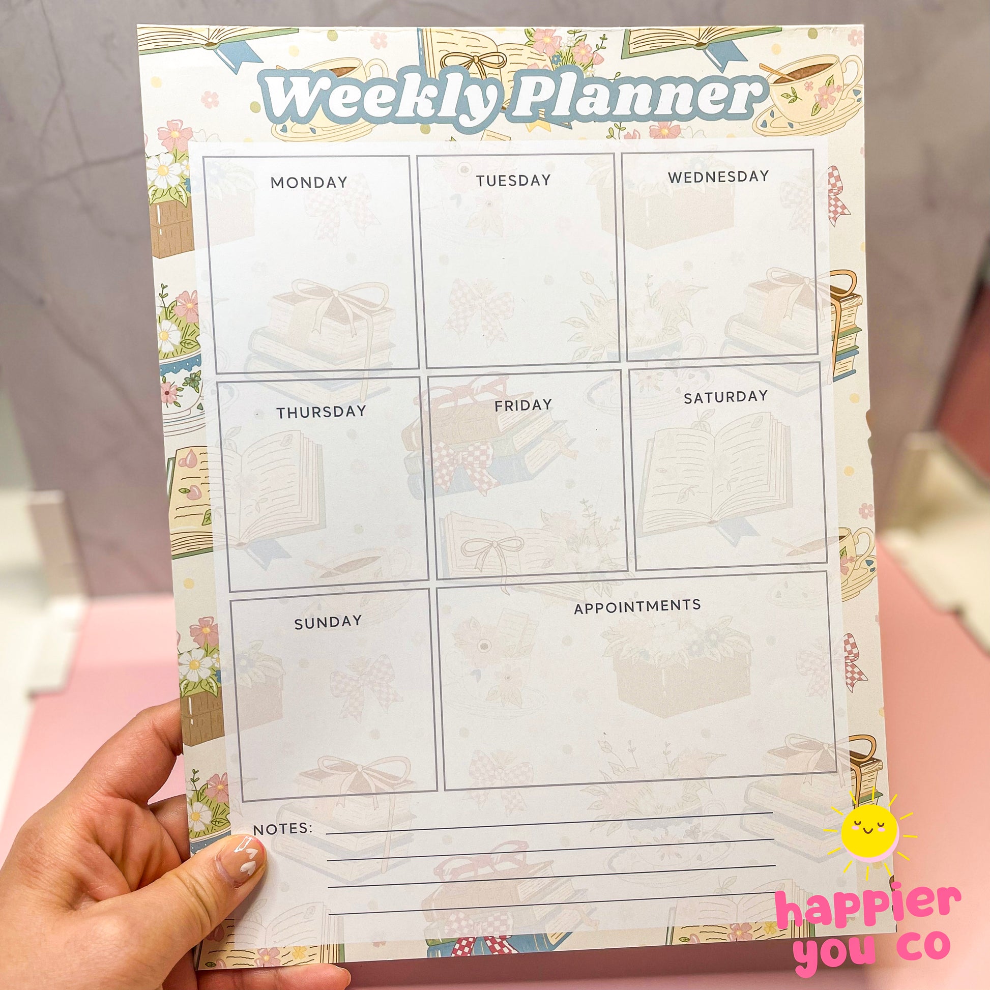 Weekly planner with bookish floral designs held by a hand, featuring the brand 'happier you co'.