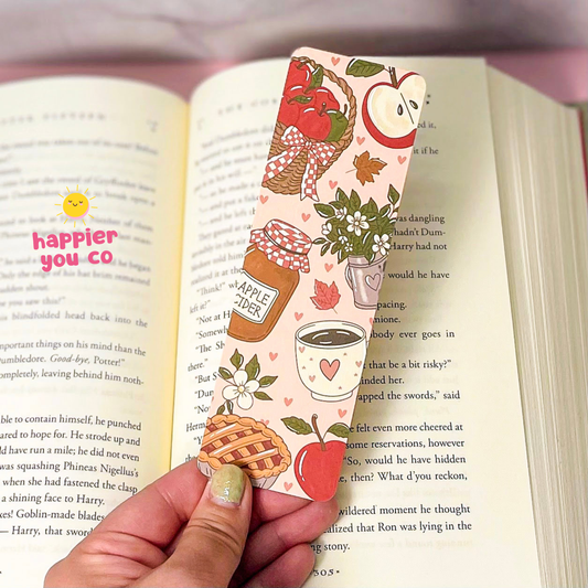 A hand with glittery green nails holds a decorative bookmark printed with autumn apple-themed illustrations, including an apple jar, apple pie, strawberries, leaves, and a teacup, over an open book.