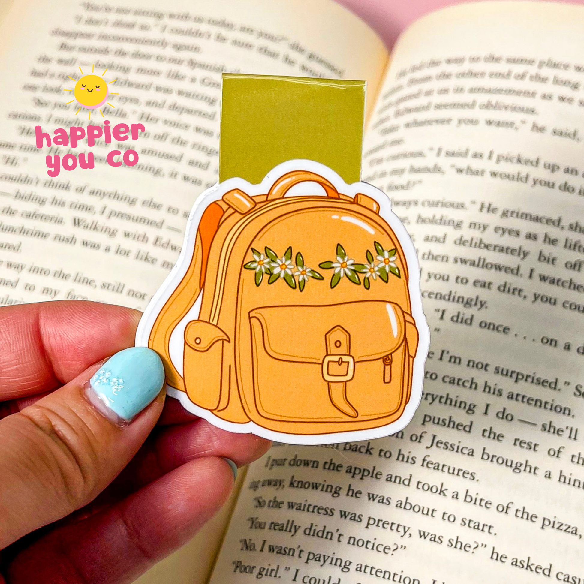 A magnetic bookmark shaped like a yellow-orange backpack with green leaves and white flowers, held by a hand over an open book. The sticker has a white border and a pink text overlay that reads 'happier you co'.