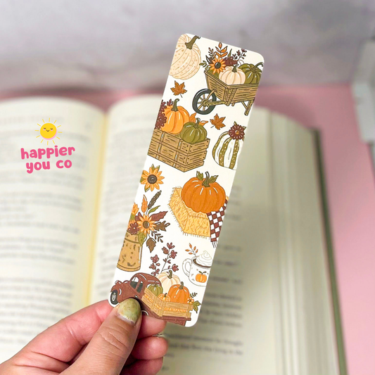 A hand holds a slim rectangular paper bookmark printed with an autumn harvest pattern featuring pumpkins, sunflowers, leaves, and a small truck, in front of an open book.