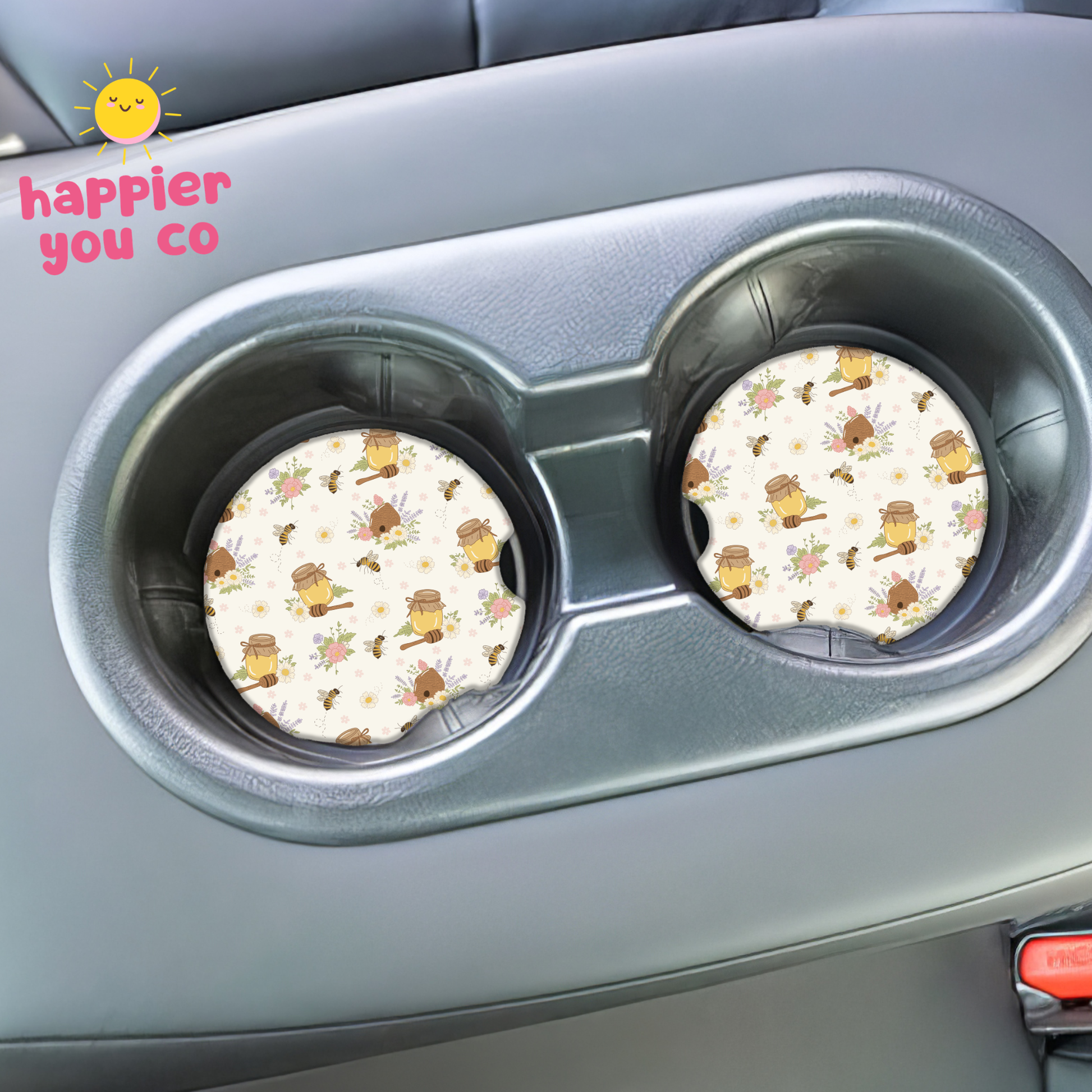 Two round coasters with a honey bee pattern in a car cup holder, branded 'happier you co'.