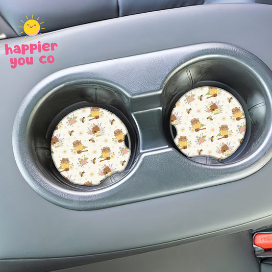 Two round coasters with a honey bee pattern in a car cup holder, branded 'happier you co'.