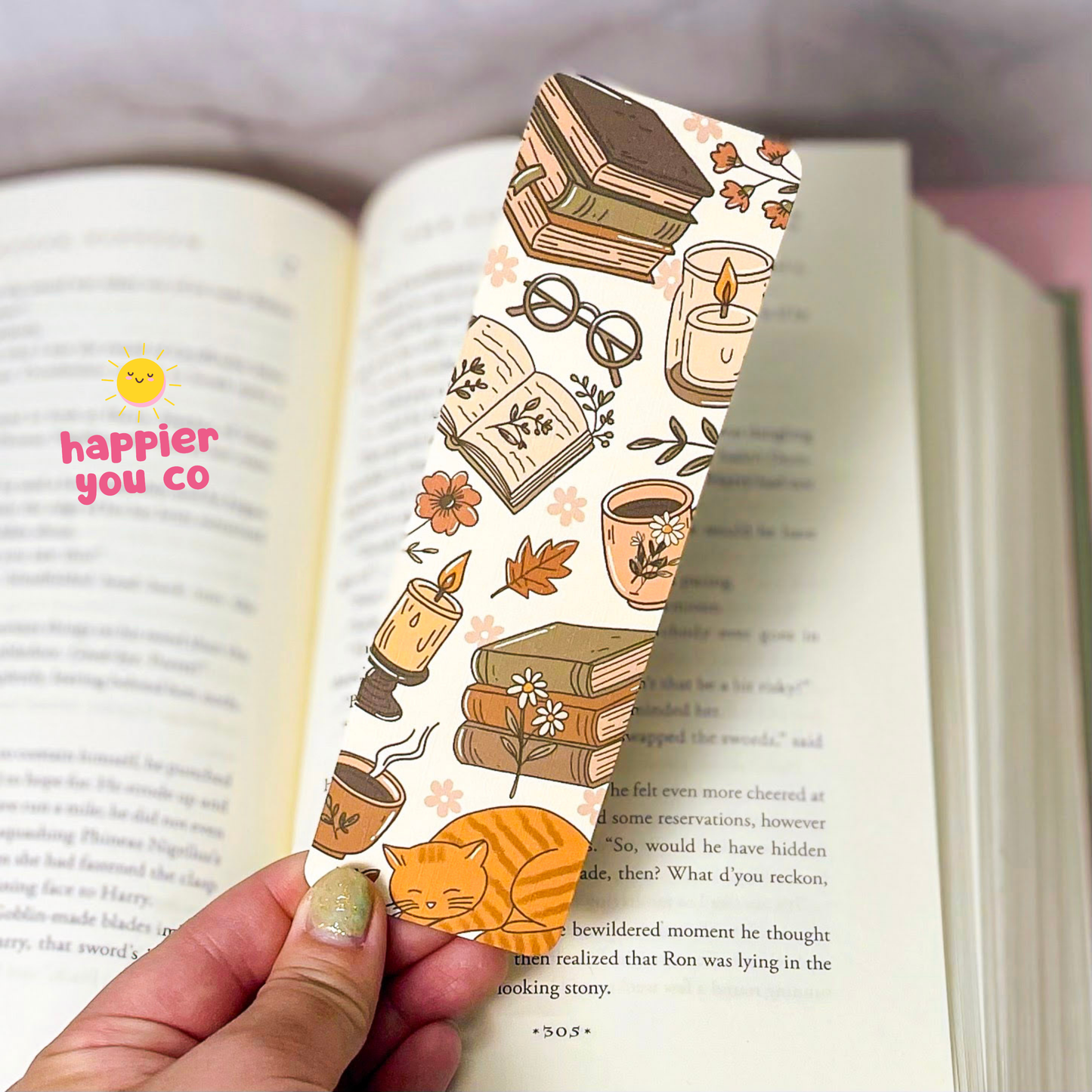 A hand holds a cream-colored paper bookmark printed with illustrations of stacked books, glasses, a candle, a mug, flowers, leaves, and an orange cat. The bookmark is positioned over an open book with printed text.