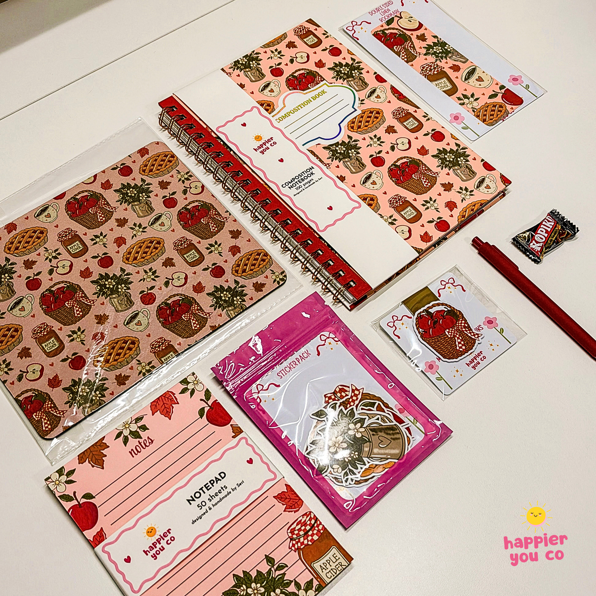 A flat-lay collection of cute, strawberry-themed stationery items on a white surface, including patterned notebooks, a notepad, sticker sheets, a pink packaged sticker pack, and a red pen.